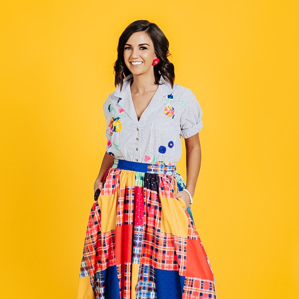 Sarah Chapman Bacerra, smiling, in an embroidered striped shirt and a colorful patchwork skirt against a yellow background